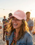 Woman wearing a pink cap with I bring the vibes on a beach with friends around her.