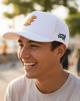 Man wearing a white cap with a dinosaur logo, smiling outdoors.