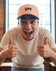 Man wearing a cap with 'yes' text, giving thumbs up in an indoor setting