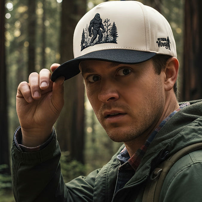Man wearing a cap with a Bigfoot design in a forest setting