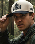 Man wearing a cap with a Bigfoot design in a forest setting