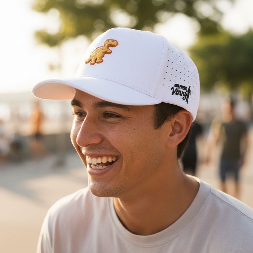Man wearing a white cap with a dinosaur logo, smiling outdoors.