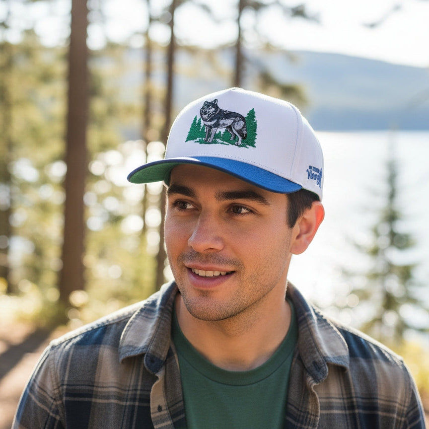 Man wearing a cap with a wolf design outdoors near trees and water