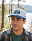 Man wearing a cap with a wolf design outdoors near trees and water