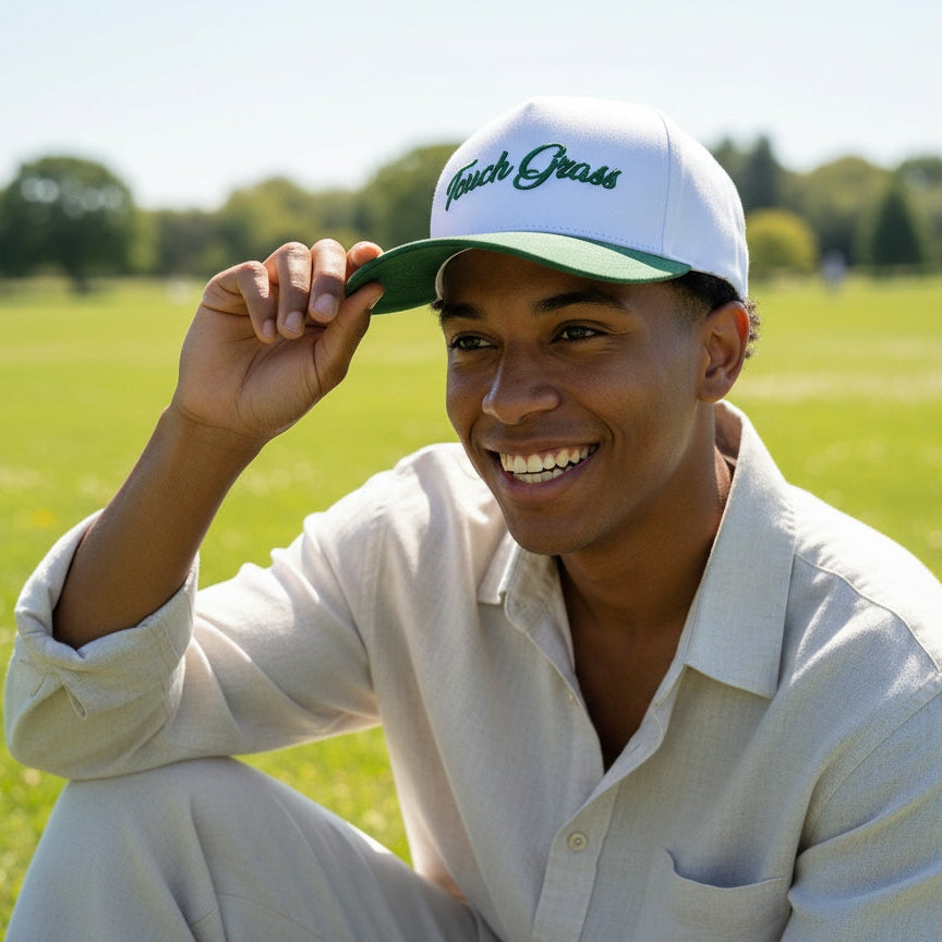 Person wearing a white cap with green brim and logo, sitting outdoors on grass.