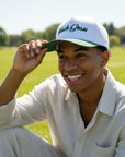 Person wearing a white cap with green brim and logo, sitting outdoors on grass.