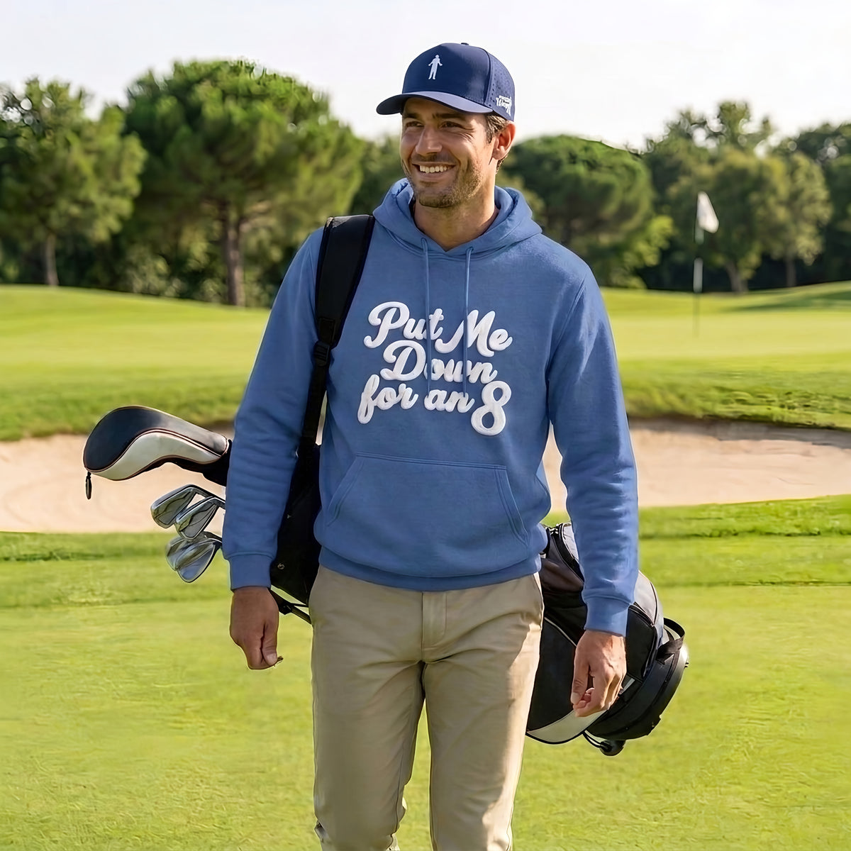 Golfer carrying his bag on the course wearing the blue Put Me Down for an 8 hoodie and a navy Our Friend Vinny cap